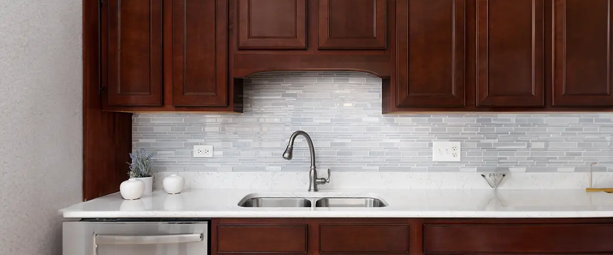 A kitchen faucet detail with a glass tiled backsplash, dark wood cabinets, and a white marble countertop.