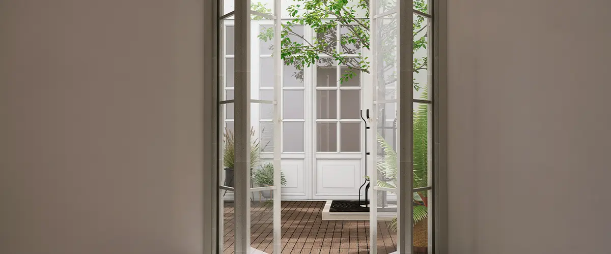 Open folding door with glass panel to courtyard with wooden floor, white exterior house and tree in sunlight