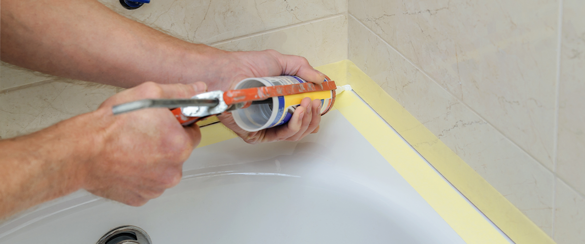 Applying silicone bathroom caulk around a bathtub edge with caulking gun for bathroom maintenance and waterproofing.