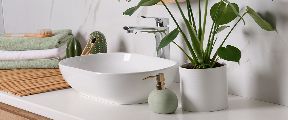 Modern bathroom with white vessel sink, green plant, round mirror, and neatly stacked towels on a wooden vanity.