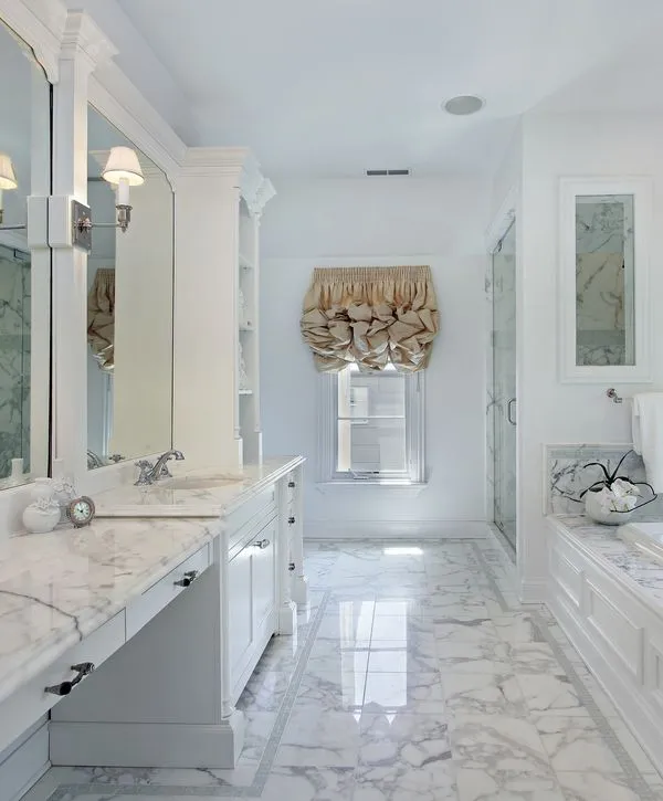 A brightly lit, long, luxury master bathroom featuring white marble tile flooring and countertops, double vanity sinks, extensive white cabinetry, and a gold-colored Roman shade above a small window, showcasing the results of expert bathroom remodeling.