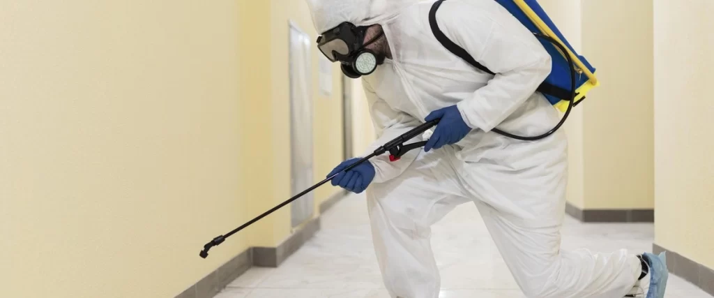 A professional mold exterminator in a white protective suit, gas mask, and blue gloves uses a long spray wand connected to a backpack sprayer to treat a wall near the floor, performing thorough mold remediation