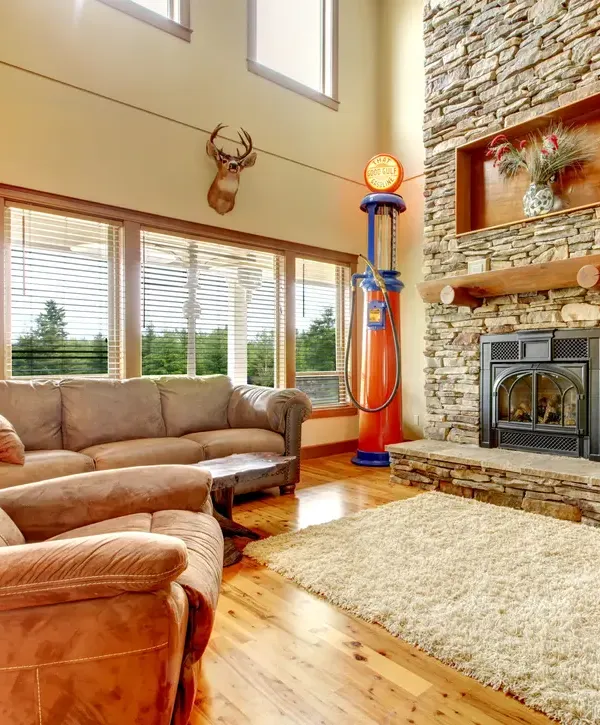 A cozy rustic living room features a prominent stone fireplace with a wooden mantel, a vintage gas pump replica, comfortable brown leather sofas, light wood flooring, and a large area rug, all illuminated by natural light from ample windows.