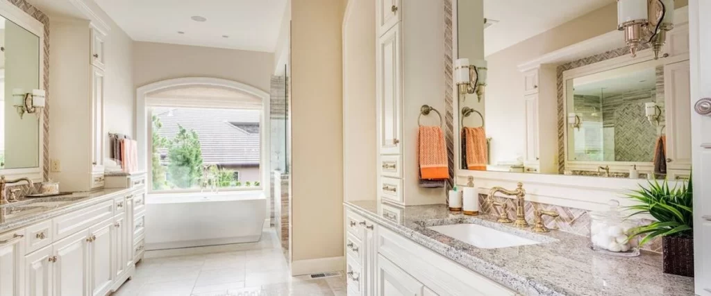 A large, bright bathroom with two separate vanities featuring white cabinetry and granite countertops, leading to a recessed archway where a modern, freestanding white soaking tub is positioned beneath a large window.