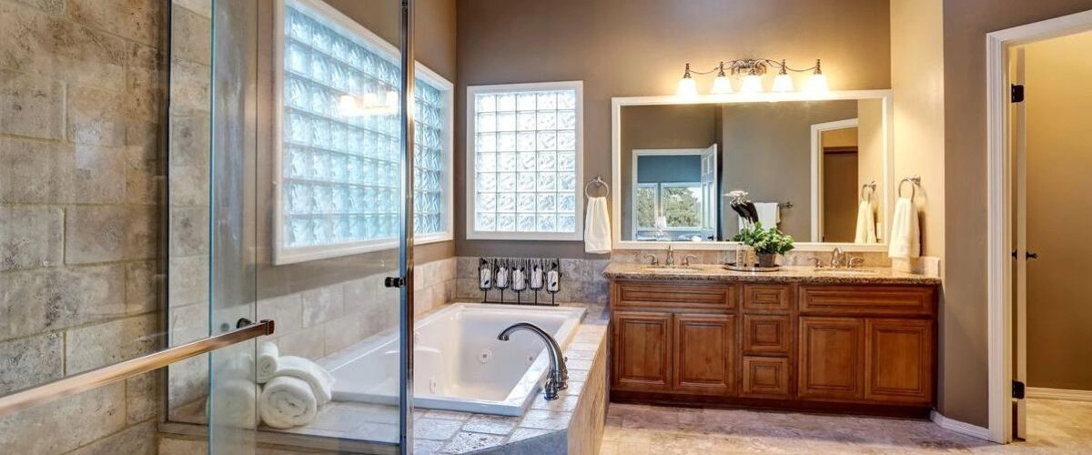 A cozy, warm-toned bathroom featuring a wood double-sink vanity with a granite countertop, a sunken jacuzzi tub, glass block windows for privacy, and a walk-in shower with large tiled walls.