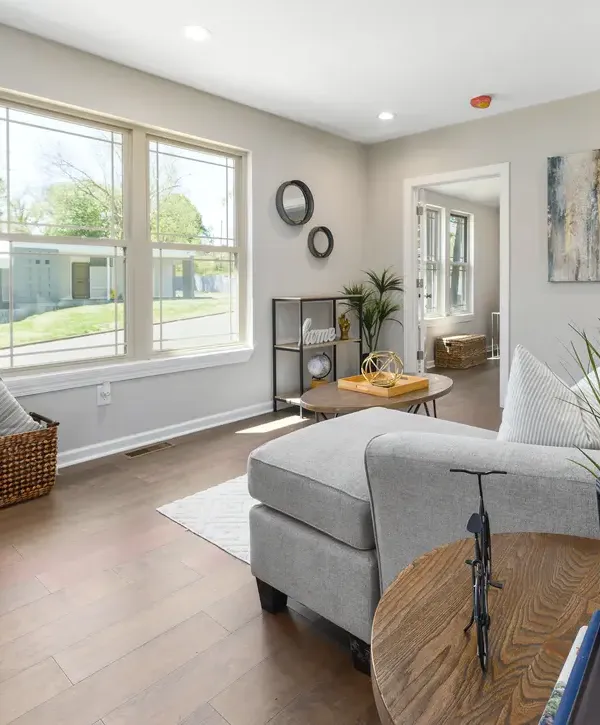 A contemporary living area resulting from a home remodeling in Burke VA, showcasing large windows that flood the room with natural light, light gray walls, a sectional sofa, modern side tables, and dark wood flooring.