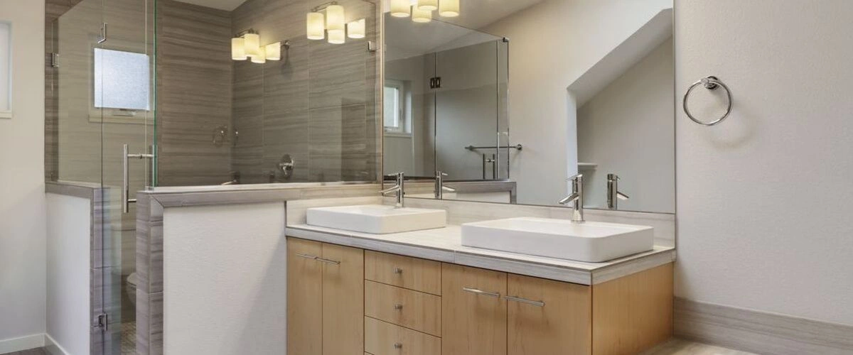 A contemporary bathroom renovation with a floating light wood vanity, dual white vessel sinks, a large mirror, and a walk-in shower enclosed by glass and tiled with horizontal gray stone.