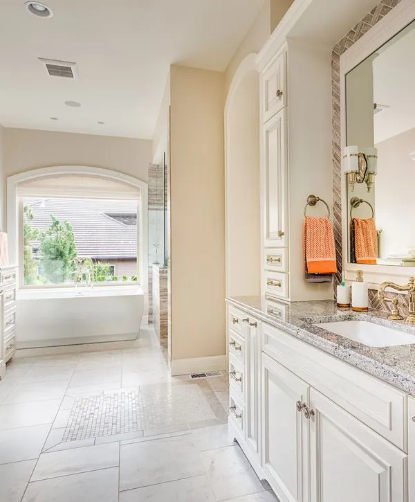 An elegant, light-filled bathroom remodeling project in Burke VA, showing white floor-to-ceiling custom cabinets, a granite vanity countertop, and a freestanding bathtub visible through an archway, illuminated by natural light.