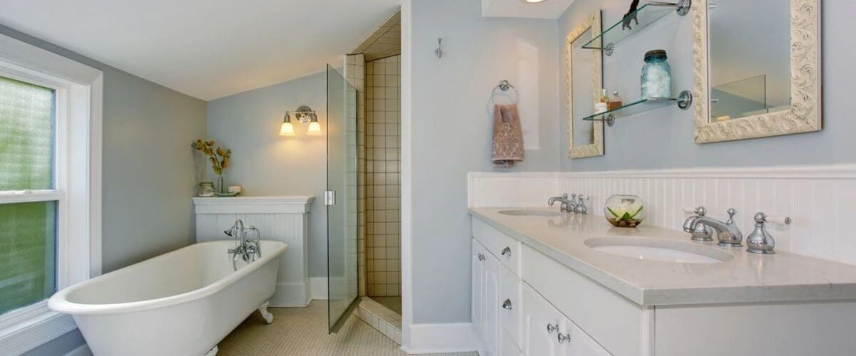 A charming bathroom with light blue walls, featuring a white clawfoot bathtub, wainscoting, an enclosed glass shower, and a white double vanity with chrome faucets and a glass display shelf.