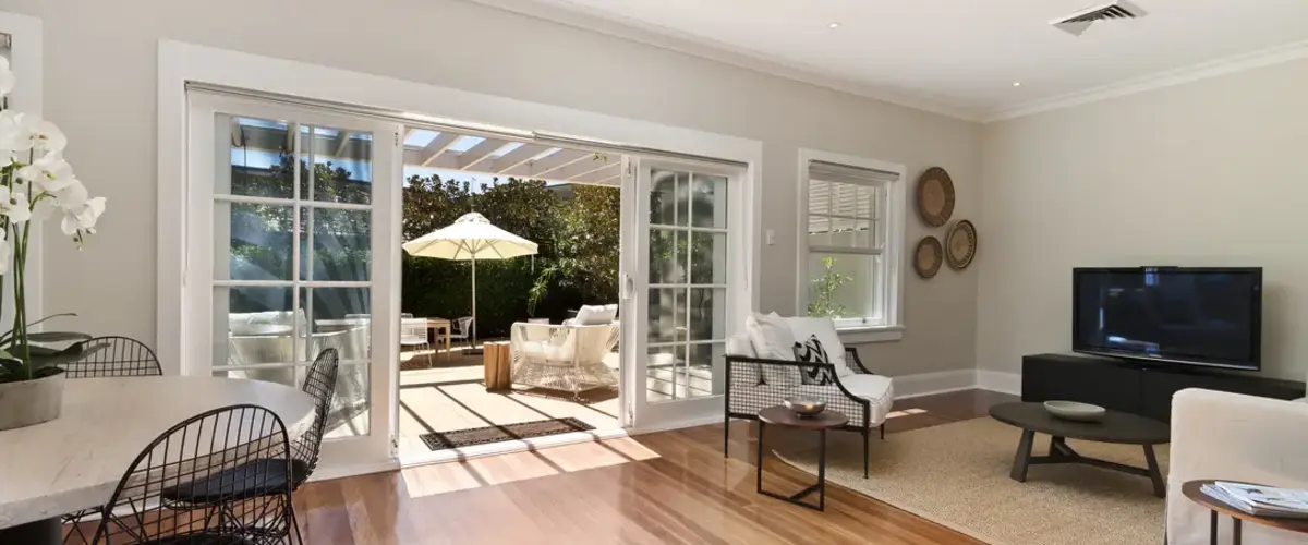 An open-concept living and dining area featuring polished hardwood floors, modern furniture, and large, white-framed sliding glass doors leading out to a sunlit outdoor patio deck with seating and a large white umbrella.