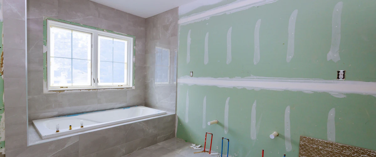 Under construction bathroom new fixtures area showing a white built-in tub, grey wall tiles, and exposed plumbing on green moisture-resistant drywall.
