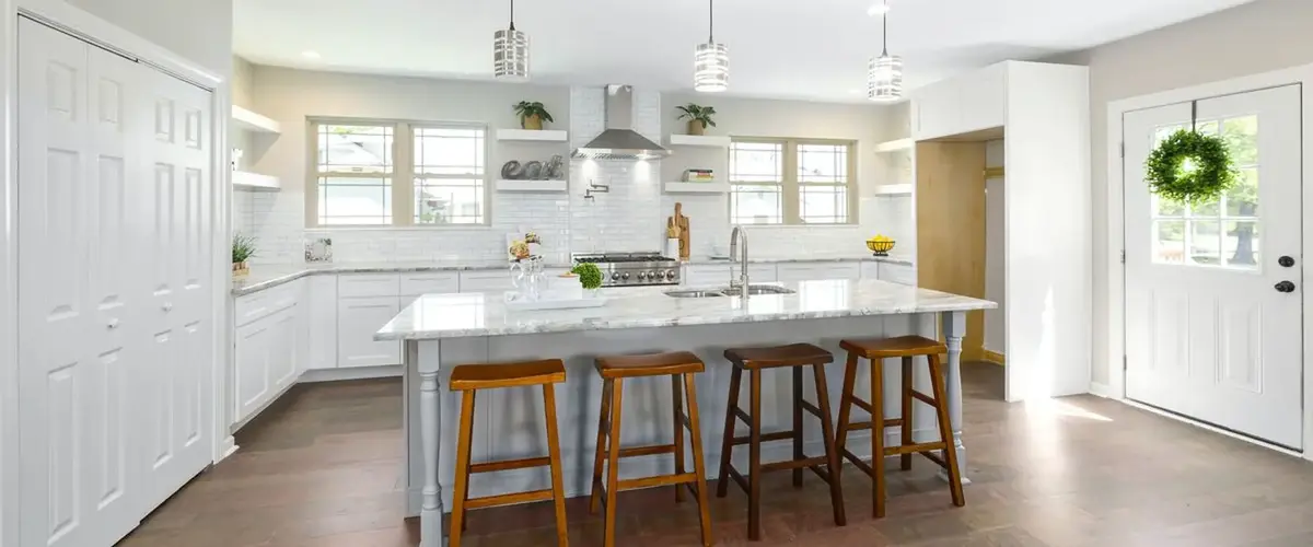 A spacious and bright white kitchen with an expansive central island, four wooden bar stools, white subway tile backsplash, and decorative greenery.