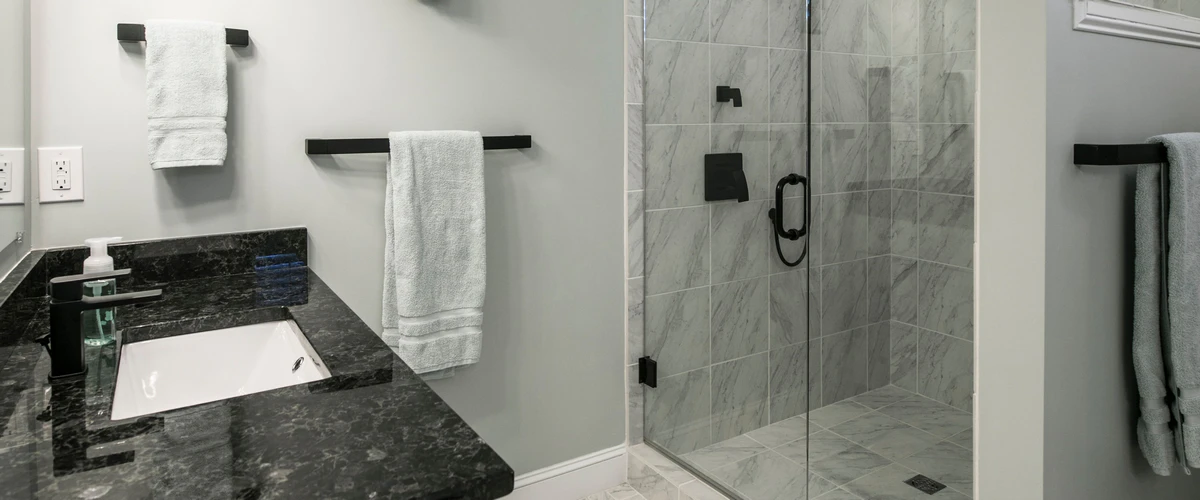 A contemporary bathroom interior showcasing a black granite vanity countertop with a white undermount sink and a glass-enclosed marble shower.