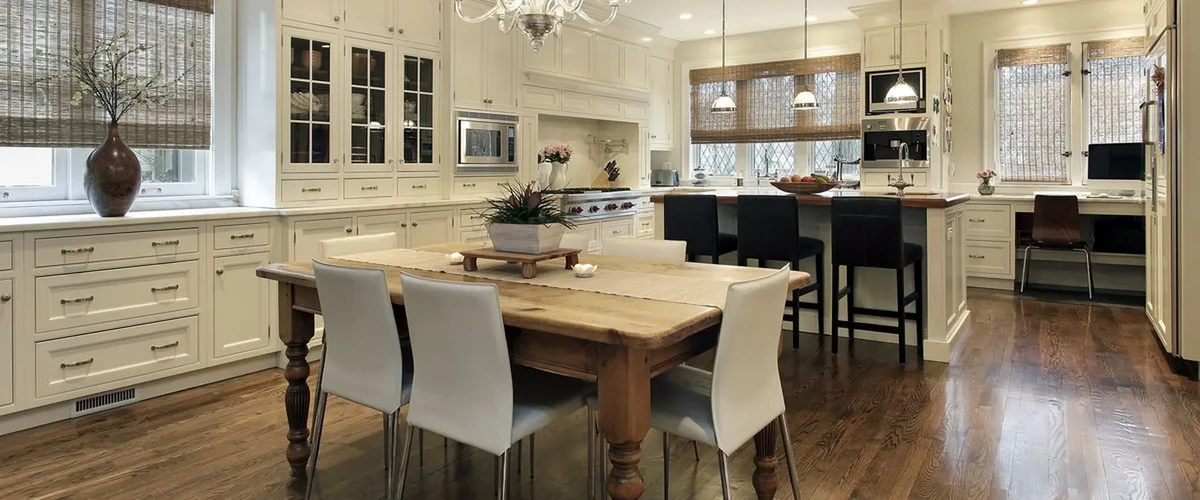 A spacious country-style kitchen with cream-colored built-in cabinets, a rustic wooden dining table, dark wood flooring, and large windows with woven shades.