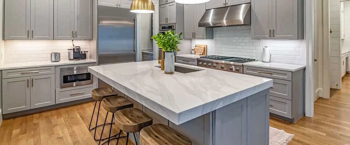 A modern kitchen featuring elegant grey cabinetry, a large white marble waterfall island with wooden bar stools, stainless steel appliances, and warm hardwood flooring.