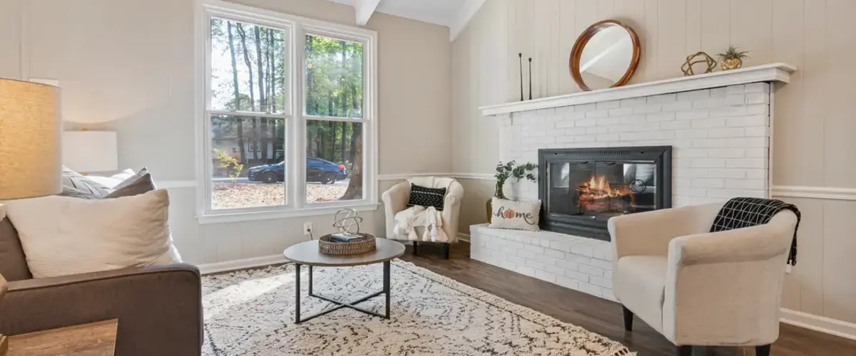 A cozy home living room featuring a white painted brick fireplace with a burning fire, two cream-colored armchairs, a patterned area rug, and a large window overlooking trees.