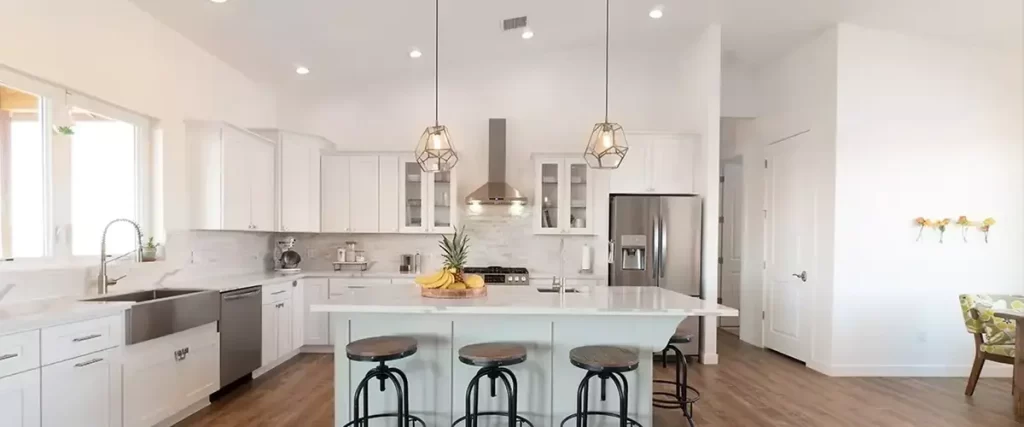 A bright and airy modern kitchen featuring white shaker cabinets, a large marble-top island with three industrial black swivel stools, and sleek stainless steel appliances.
