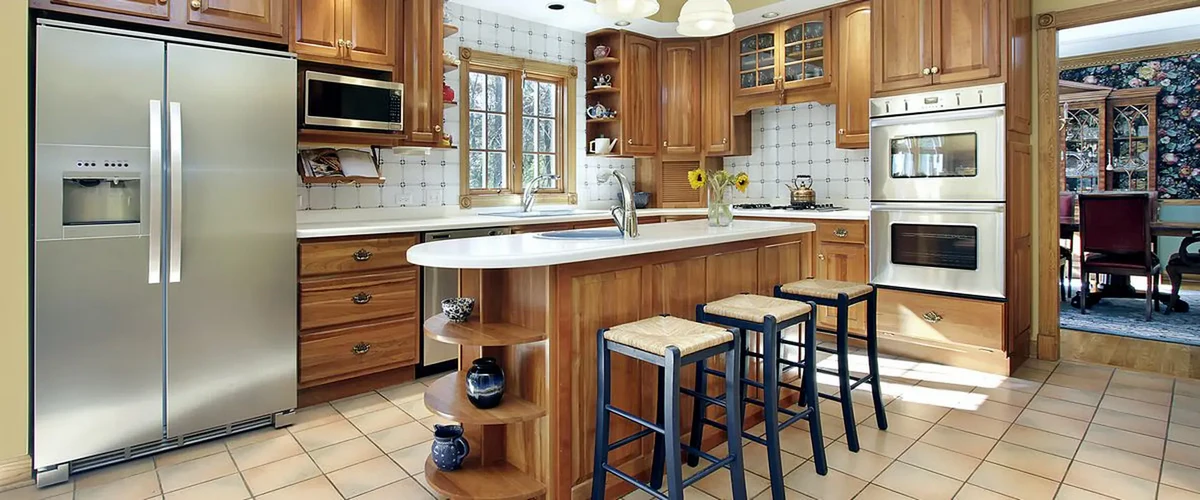 A traditional kitchen featuring warm oak wood cabinetry, a large double-door stainless steel refrigerator, and a functional center island with wicker-seat bar stools on a tiled floor.