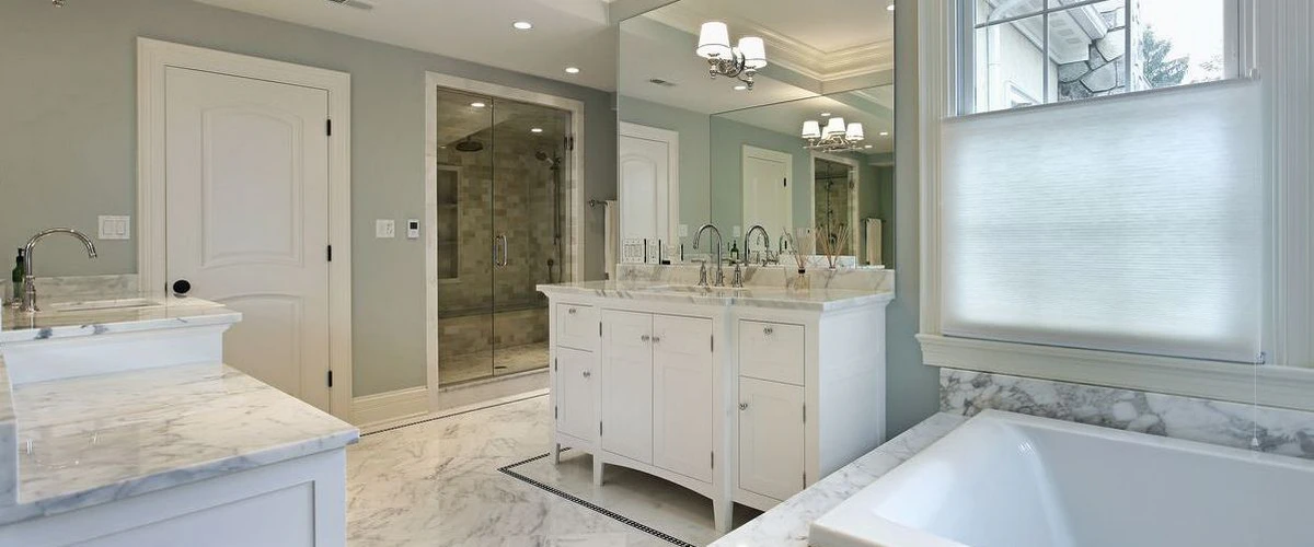 An elegant bathroom with a white double vanity, marble countertops, matching marble tile floors with black borders, and a walk-in glass shower.