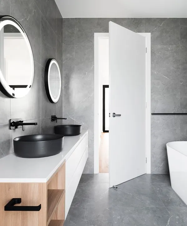 A minimalist master bathroom renovation by bathroom remodeling companies in Union Mill, VA, featuring dual black vessel sinks, backlit round mirrors, and gray marble-effect wall tiles.