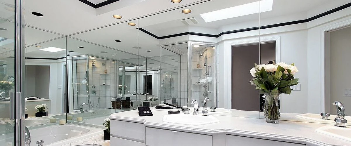 A bright and expansive white bathroom featuring wall-to-wall mirrors, a glass-enclosed shower, a soaking tub, and natural light from a ceiling skylight.