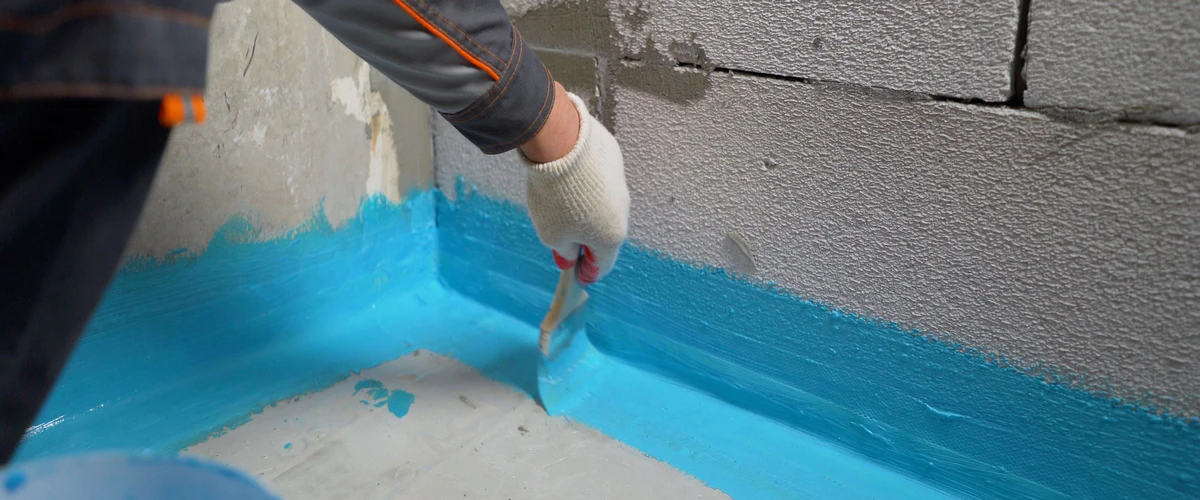 A construction worker applies a bright blue liquid waterproofing membrane to the corner joint of a concrete block wall and floor during a basement waterproofing project.