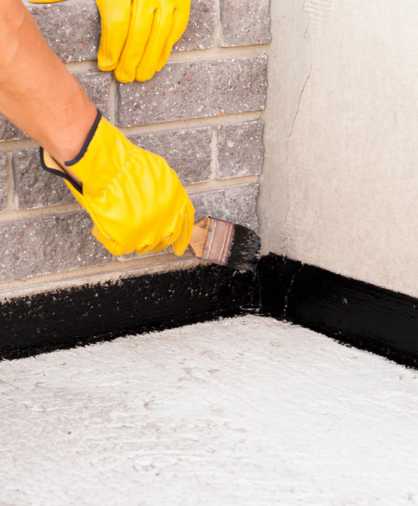 A worker wearing yellow gloves uses a brush to apply a thick black liquid membrane to the base of an interior brick wall for basement waterproofing in Centreville, VA.