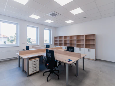 A newly finished entry-level commercial remodel featuring a bright, minimalist open office space with modular wooden desks, black ergonomic chairs, and large empty shelving units.