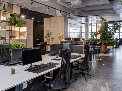 An expansive open-plan workspace showing a full commercial renovation, featuring an exposed industrial ceiling, modern desk stations with dual monitors, custom wood dividers, and abundant indoor greenery.