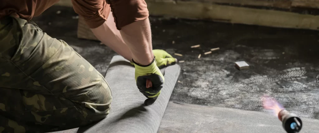 A contractor kneels to unroll a heavy-duty grey sheet membrane over a dark floor, with a lit blowtorch nearby, preparing for a basement waterproofing installation.